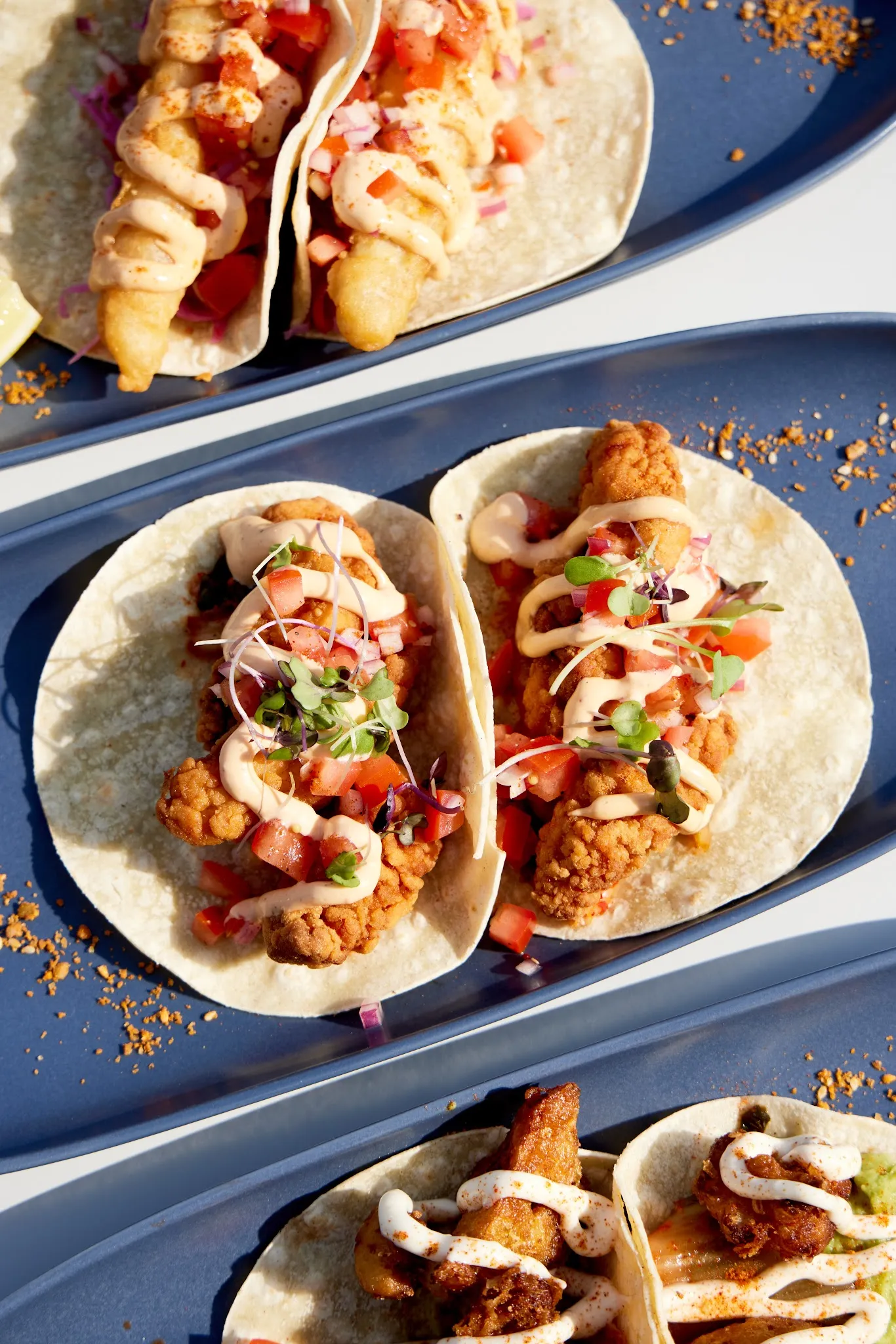 Three blue trays each holding two crispy tacos with pico de gallo and chipotle cream
