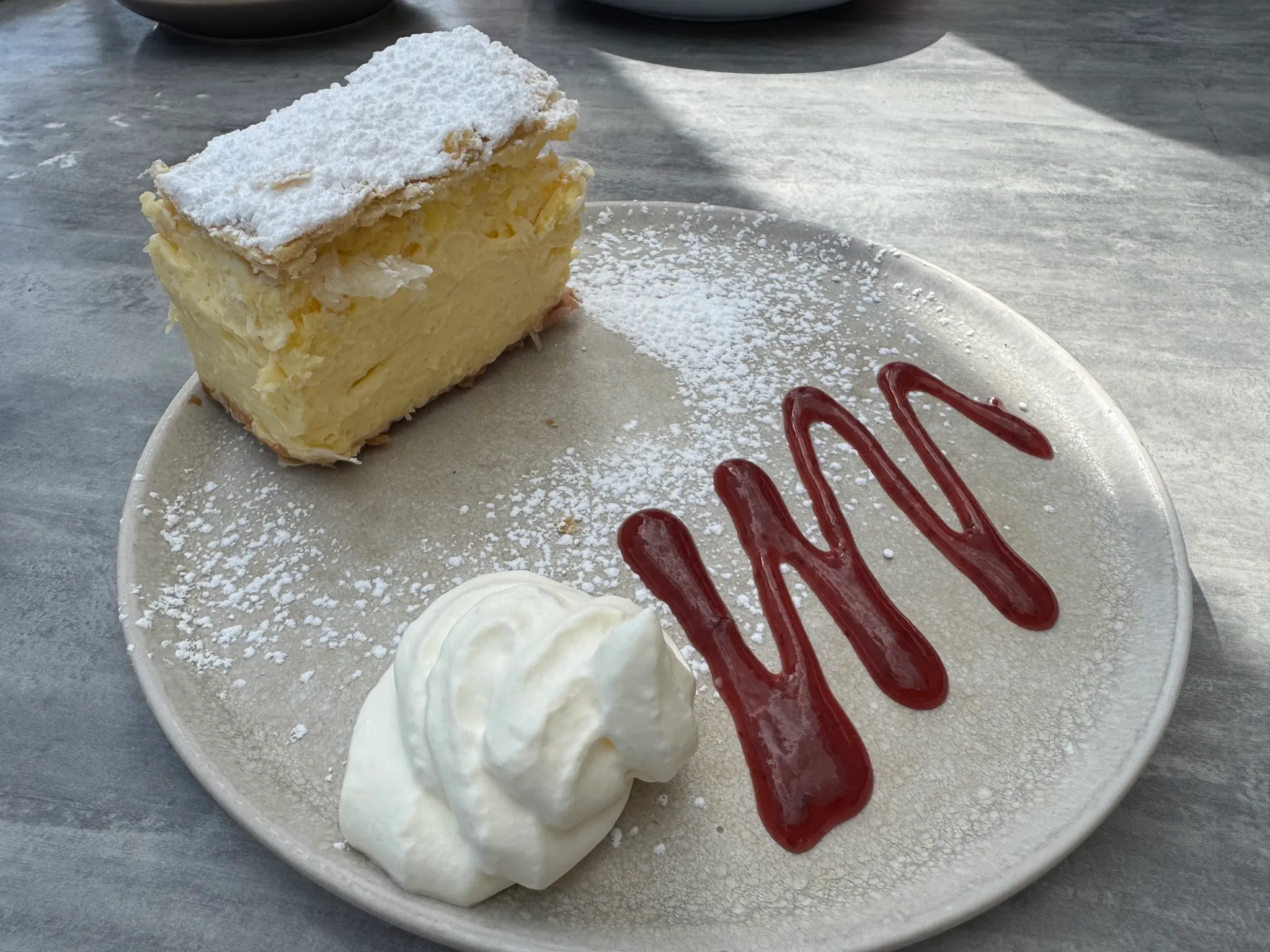 Vanilla custard slice with icing sugar on a grey plate with raspberry coulis and cream