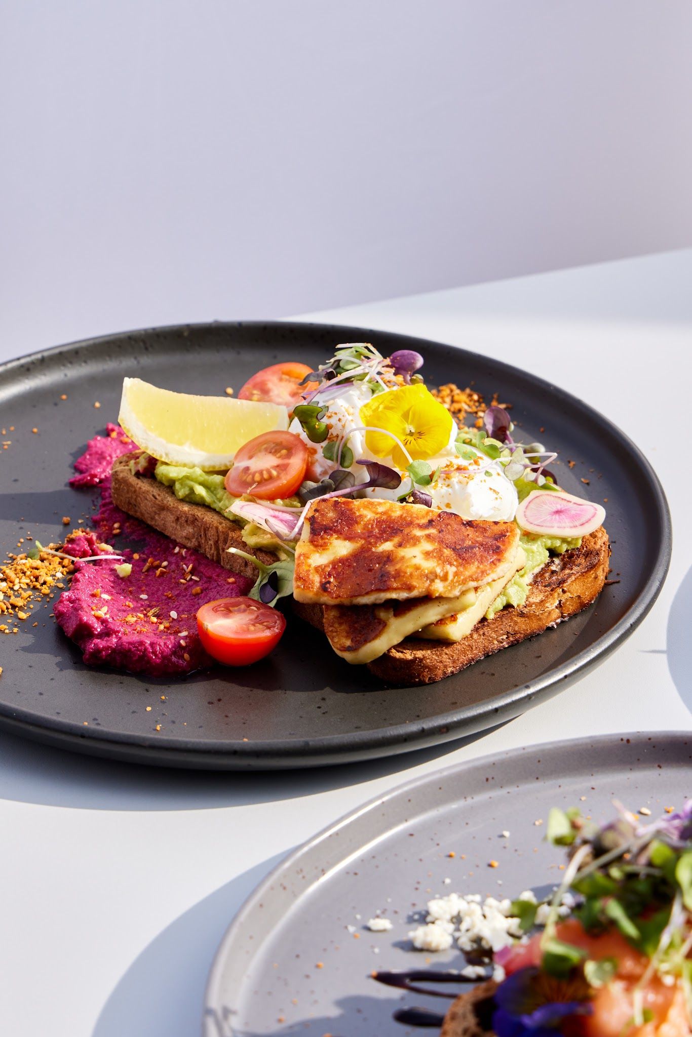 Avocado toast with halloumi, edible flowers, beetroot hummus and poached egg on dark ceramic plate