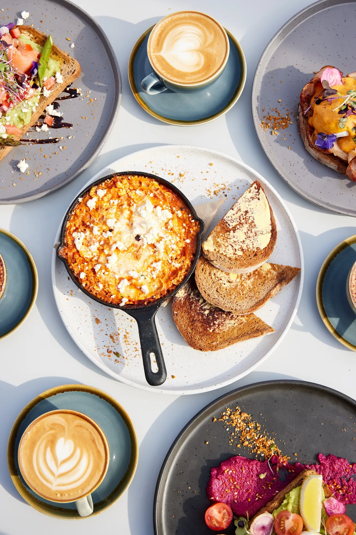 Overhead brunch spread with shakshuka, lattes and avocado toast on elegant grey tableware