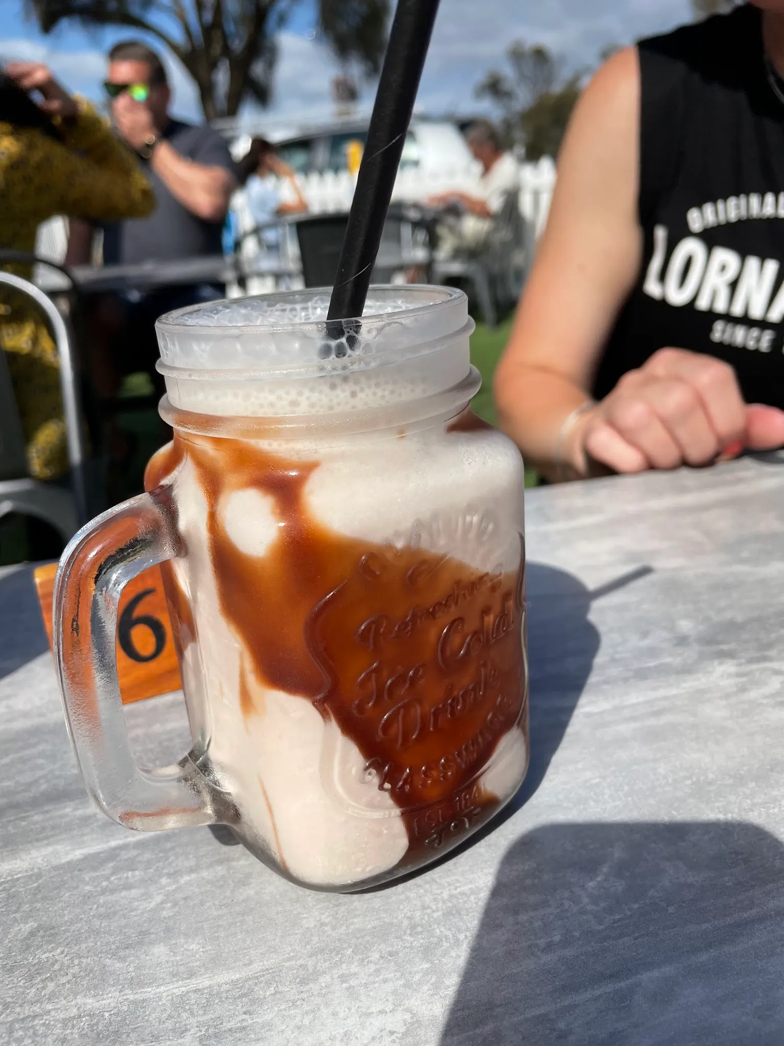 Iced coffee in a mason jar mug with a black straw, photographed outdoors with customers in background