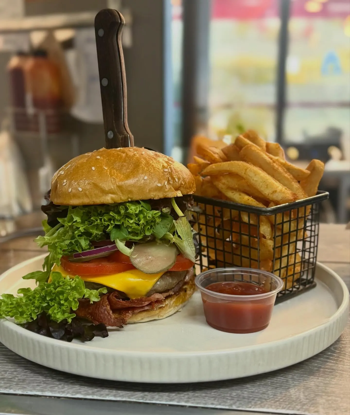 Tall gourmet burger stacked with bacon and cheese on a white tray with a steak knife through the bun